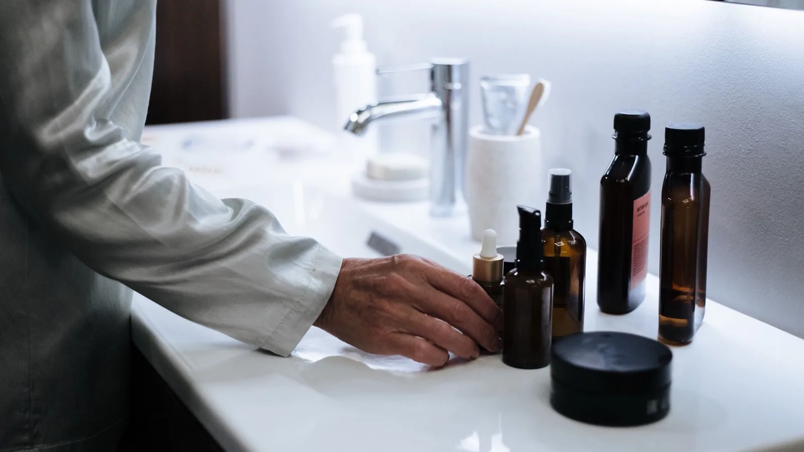 Hand reaching for a few skincare products arranged on a bathroom sink.