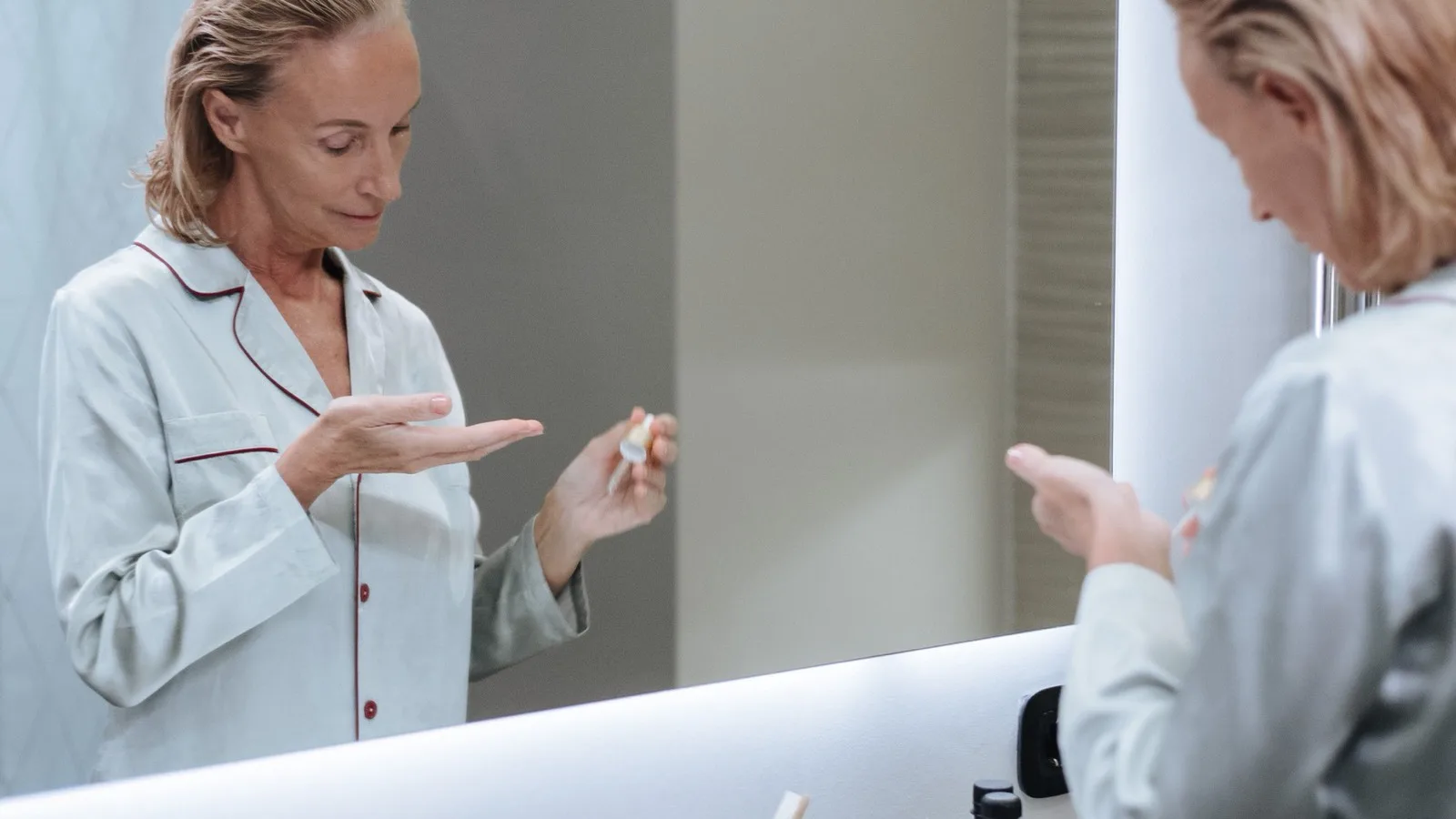 A senior woman applying serum in front of a bathroom mirror.