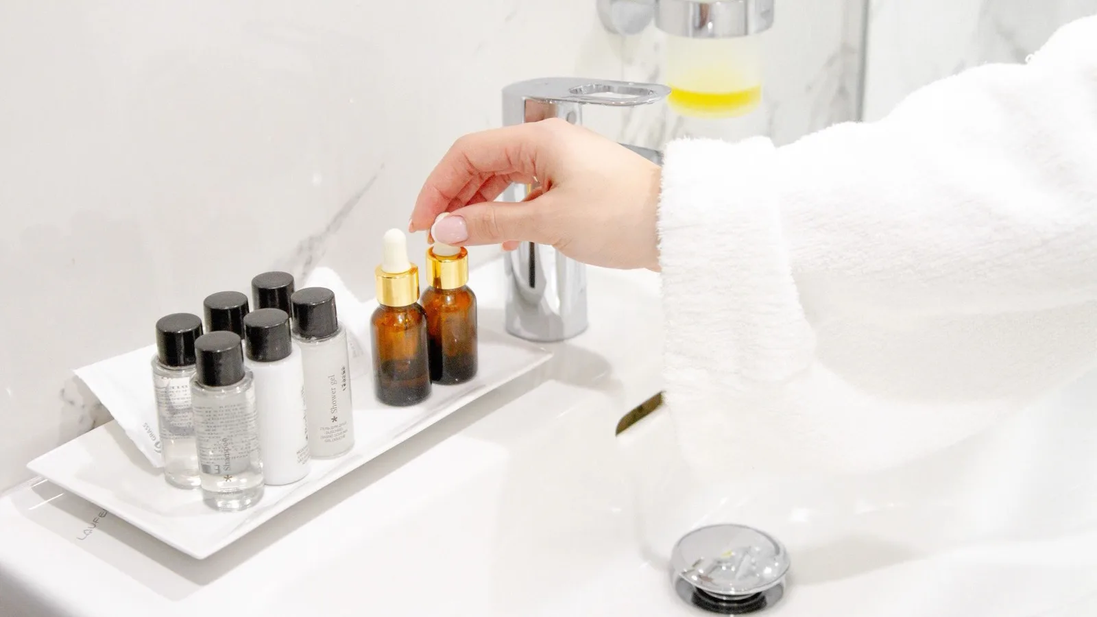 Hand reaching for a skincare serum among a few products on a bathroom sink.