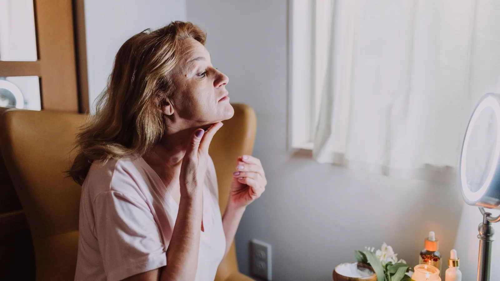 A mature woman examining her skin with skincare products nearby.