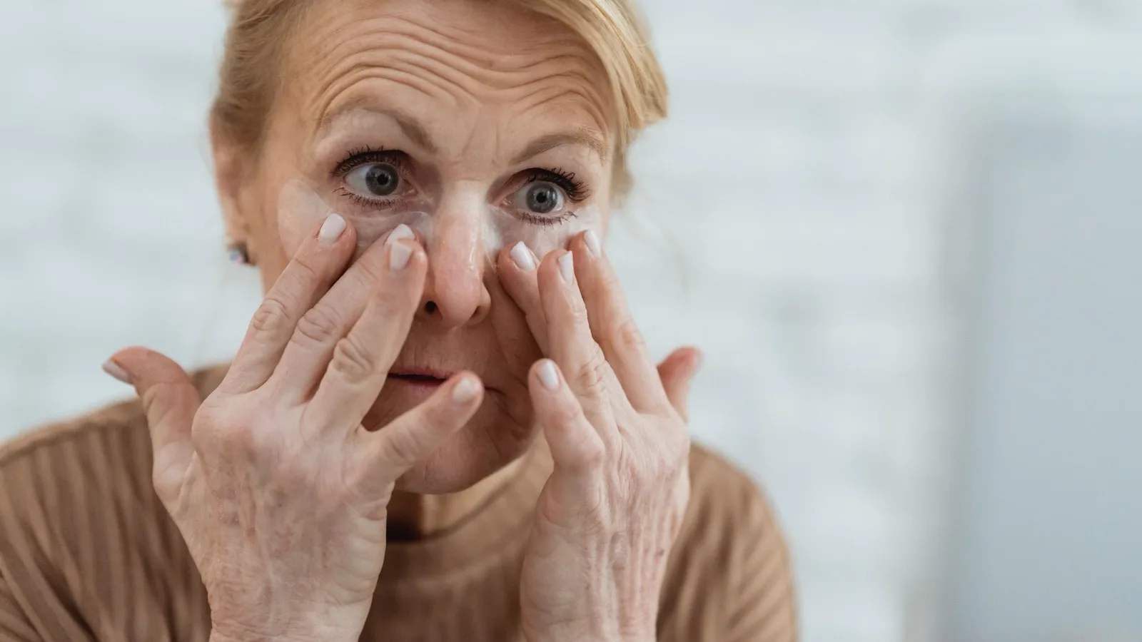 A mature woman applying hydrating eye patches in a bright bathroom.