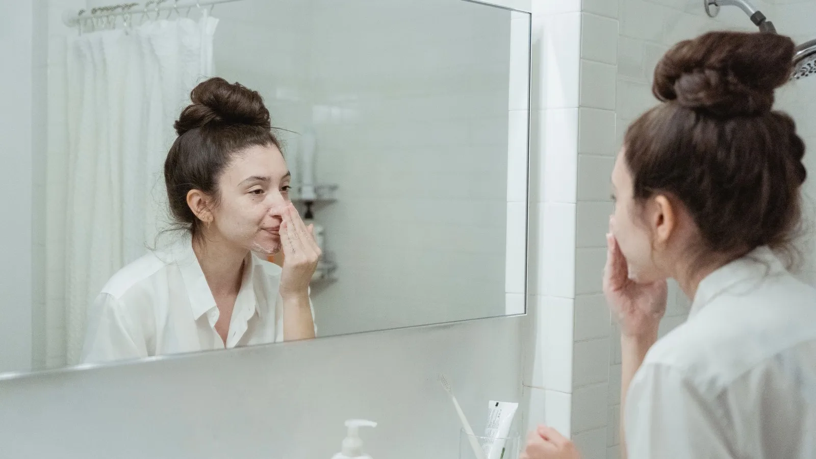 Woman applying skincare while looking in a bathroom mirror.