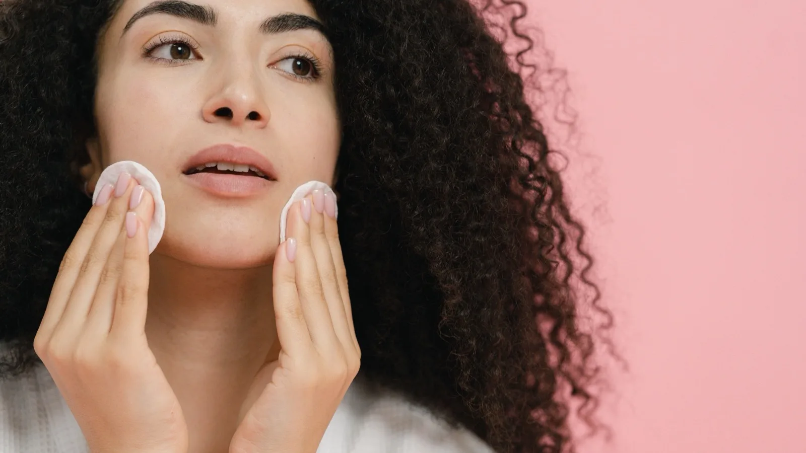 Person gently cleansing their face with a cotton pad indoors.